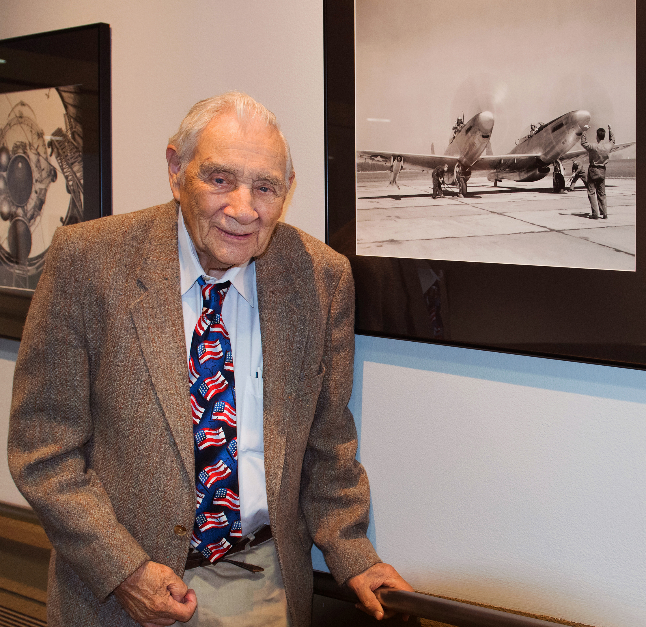 Elderly gentleman poses next to an old photograph displayed on a wall.