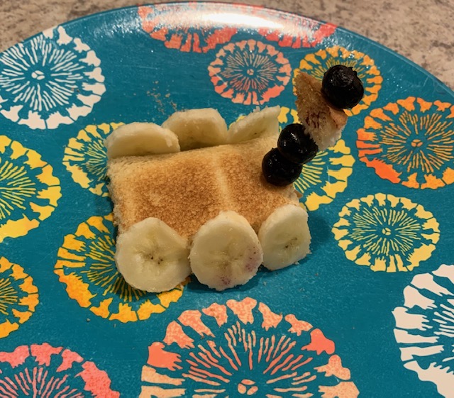 Toast with six banana slices, three on each side, representing wheels and a toothpick with blueberries, to make the toast look like a Mars rover on a blue plate with orange, white and yellow flowers.