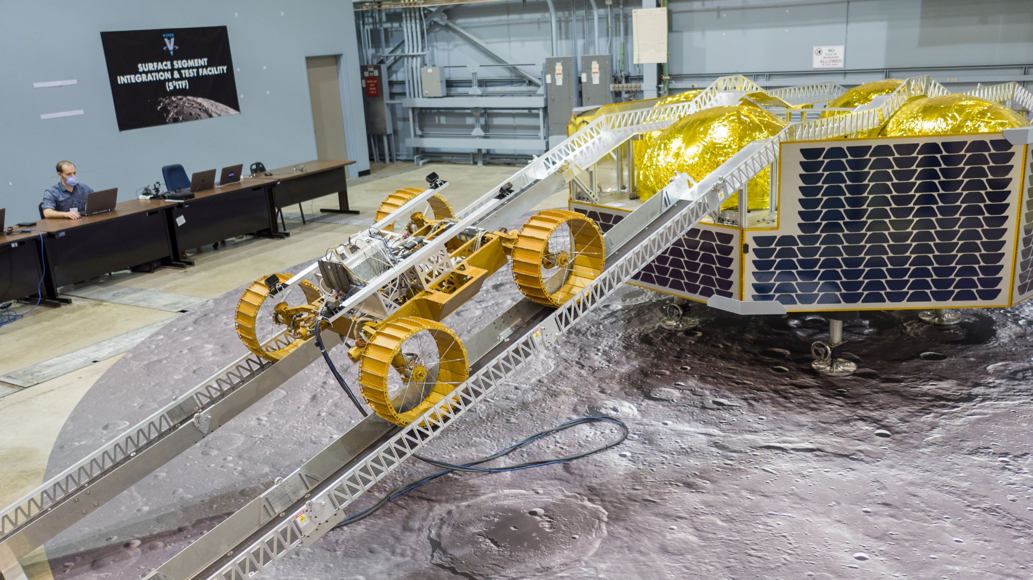 The base of a four-wheeled robotic rover on metal ramps descending from a mock-up of a lunar lander, inside a room with floor depicting the lunar surface. A person sits at a laptop behind a desk in the background.