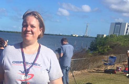 Woman with fair skin and short blonde hair wears a grey t shirt that says "OSIRIS REx" smiles in front of the ocean at Kennedy Space Center. There is a man with a camera in the background and camp chairs set up along a fence line facing the ocean. 