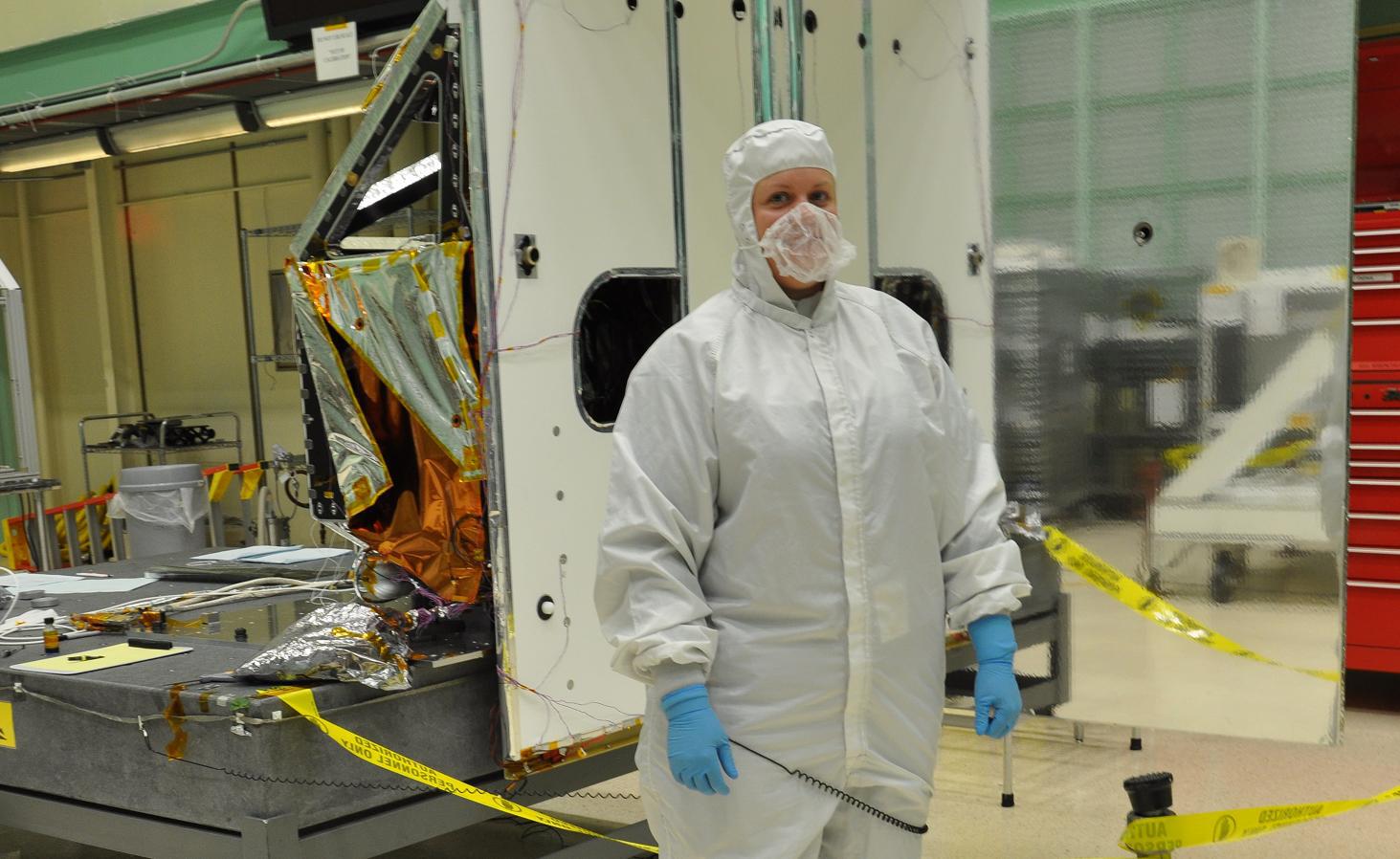 A woman wearing a white coverall suit, lightweight white mask, and blue gloves stands in a cleanroom in front of NASA/USGS' TIRS-1 instrument, a thermal sensor that measures land surface temperature. To her left, orange and silver protective sheeting hangs on a frame above a dark gray desk with a yellow clipboard and other papers sitting on top. A trash can and ladder are visible behind the desk. To the woman's right, a reflective panel mirrors the green and white walls of the cleanroom and other boxes and cabinets. On either side of her are strips of yellow caution tape.