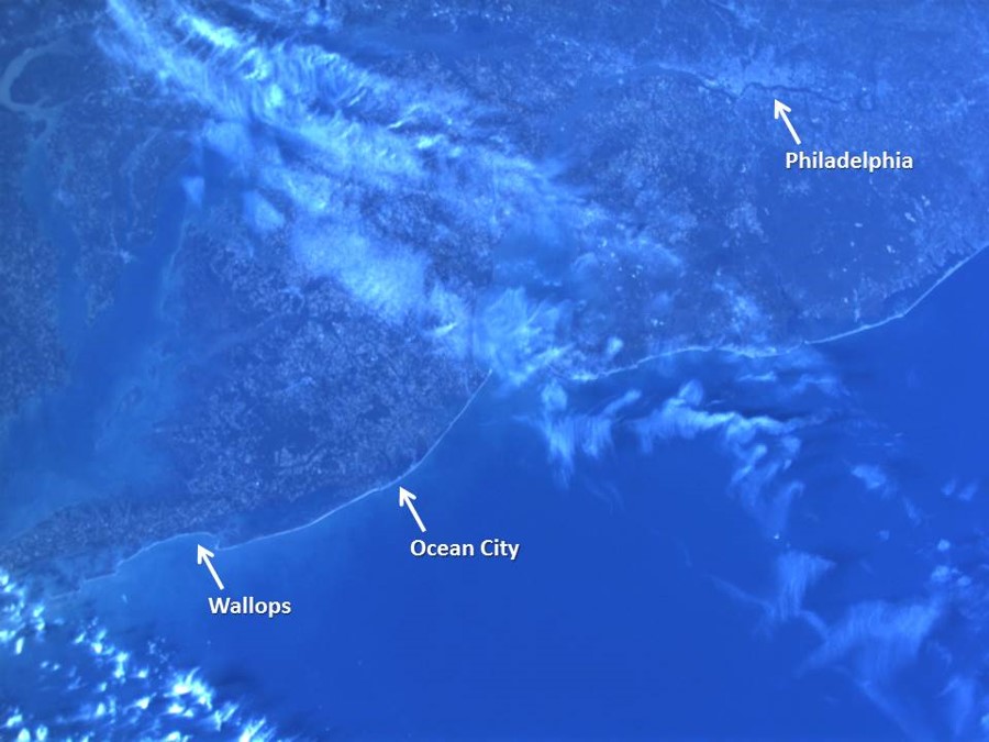 Satellite image of the coastal Maryland area. The whole image is blue. The image is divided into two triangles, diagonally from the top right to the bottom left. Land covers the top triangle. There are arrows pointing to and labeling "Wallops," "Ocean City," and "Philadelphia"  