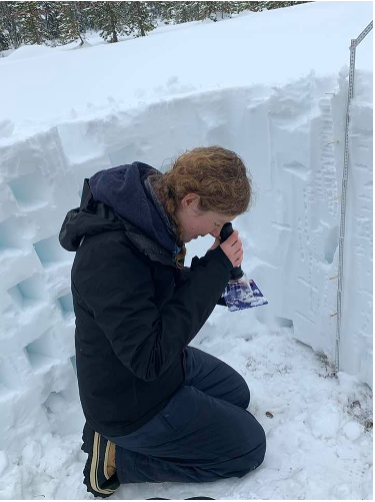 A young woman kneels in a deep pit dug in the snow, looking through a scientific instrument at a purple piece of plastic dusted with snowflakes.