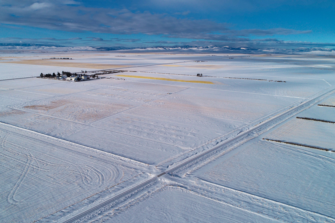 An aerial photo of snow-covered prairies, divided into large squares by intersecting roads. The fields reflect the sky in shades of light blue, white, gray, and gold where plant life shows through. A small farm is visible near the horizon, with dark evergreen trees growing around it. 