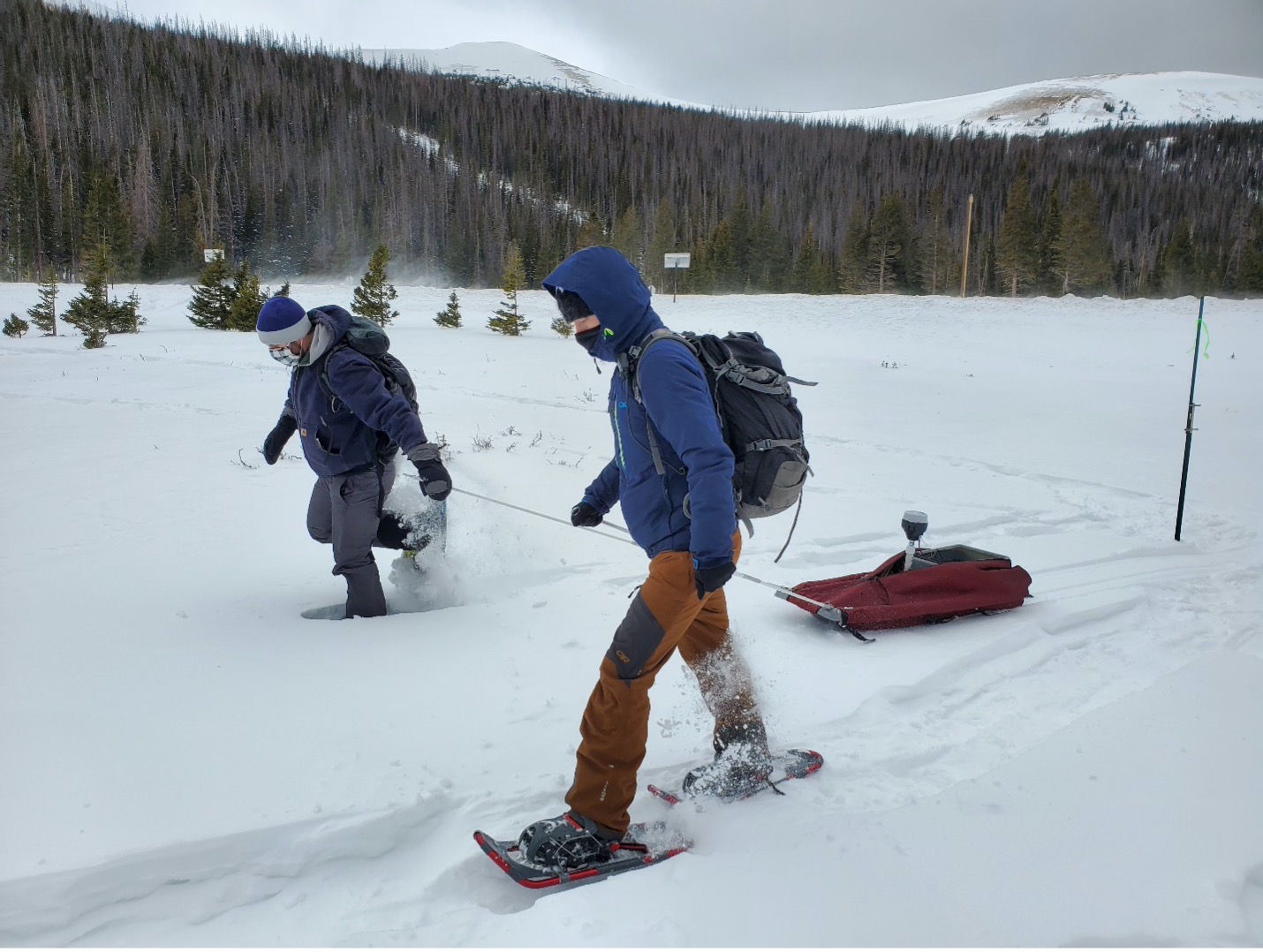 Two people in winter gear trek through calf-deep snow wearing snowshoes and pulling a small sledge behind them. The sledge is covered with a red covering and has the tube-like top of a scientific instrument sticking out of the covering. In the distance, gray-green trees slope up to snow-covered mountains and a gray sky.