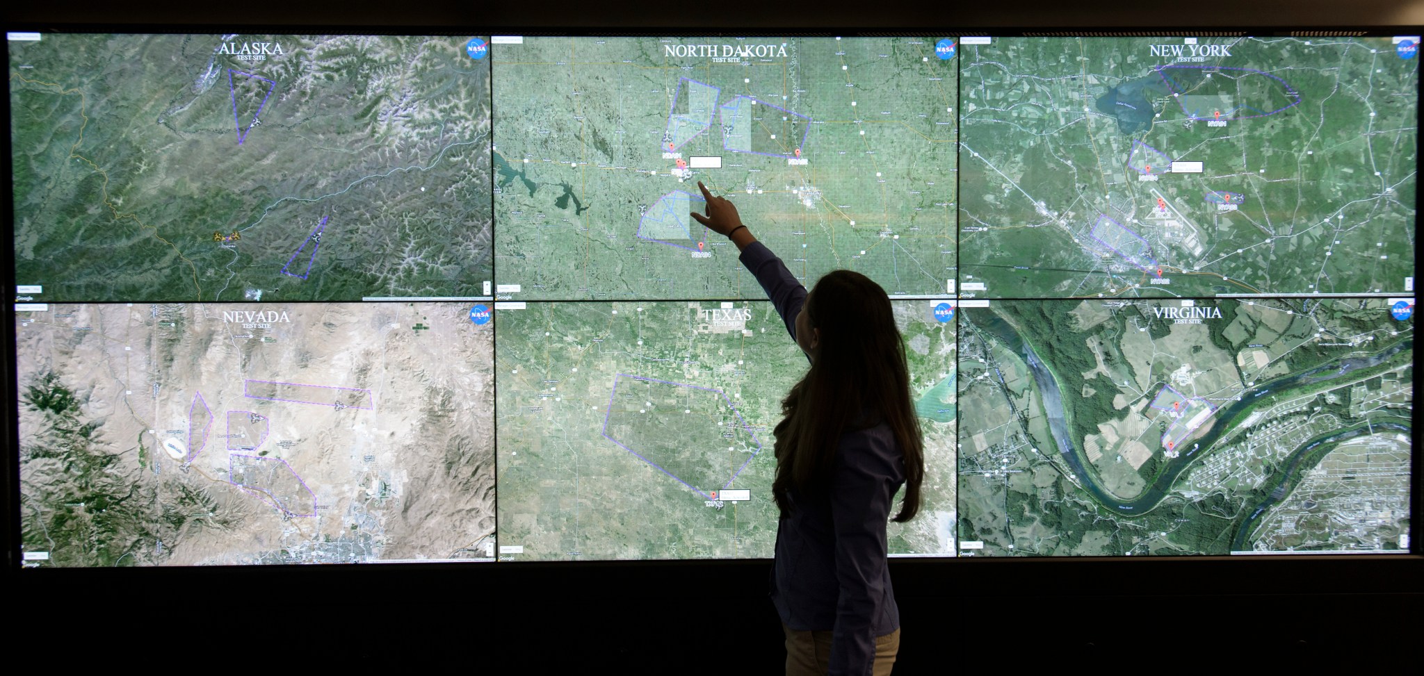 A person in silhouette, pointing up at a wall of screens displaying maps