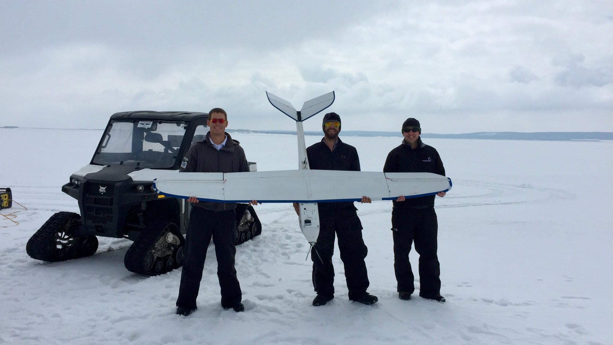 Three people in winter clothes hold a white fixed-wing drone while standing in a snow-covered landscape with a vehicle behind.