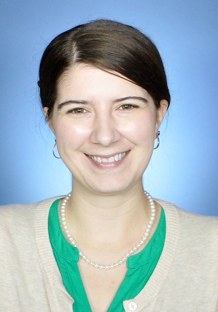 Woman with fair skin and brown hair, pulled back, wears a green shirt with a tan cardigan. She is also wearing a pearl necklace and silver hoop earrings. The background is blue. 