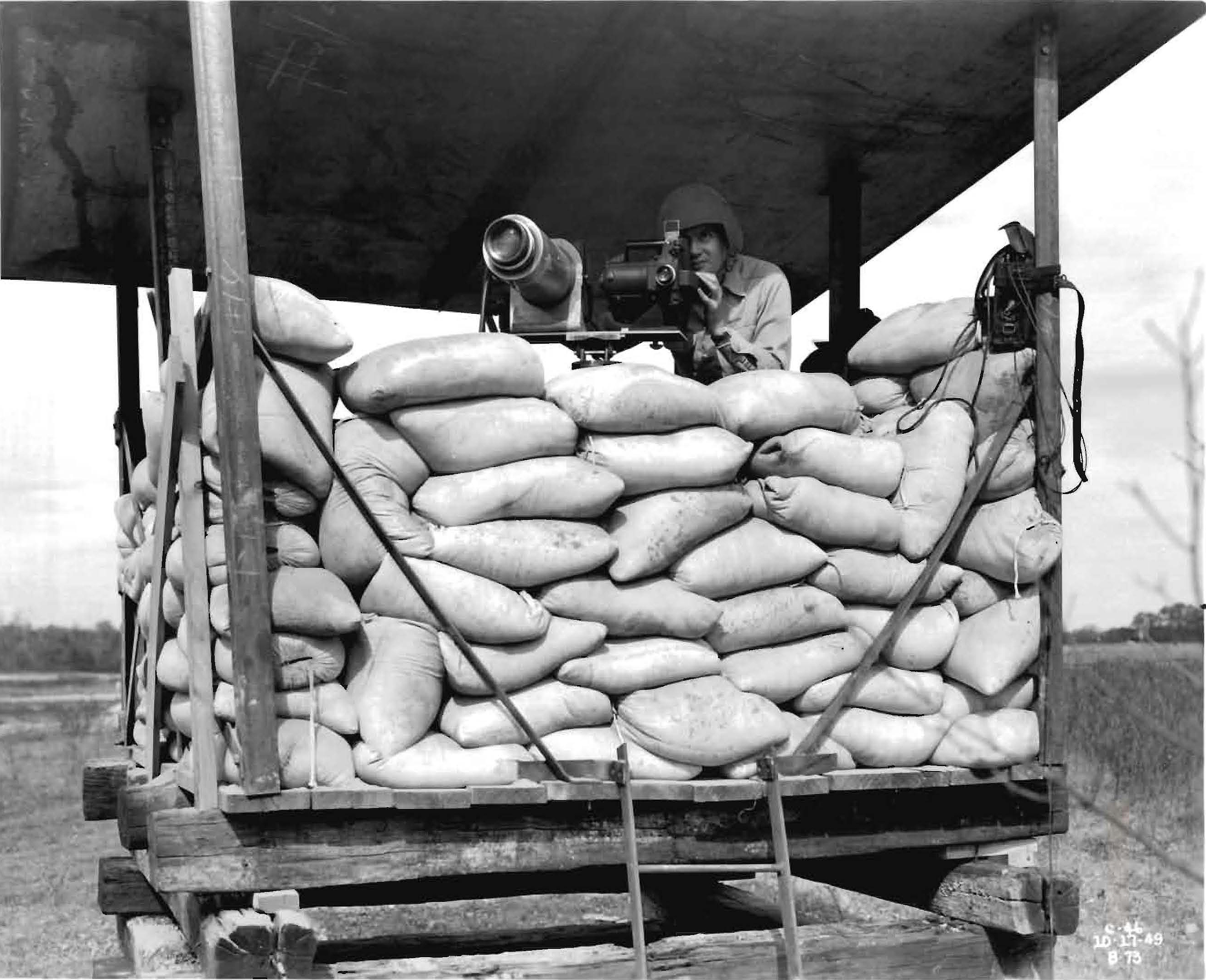 Photographer with steel helmet films an aircraft test behind sandbags on a platform.