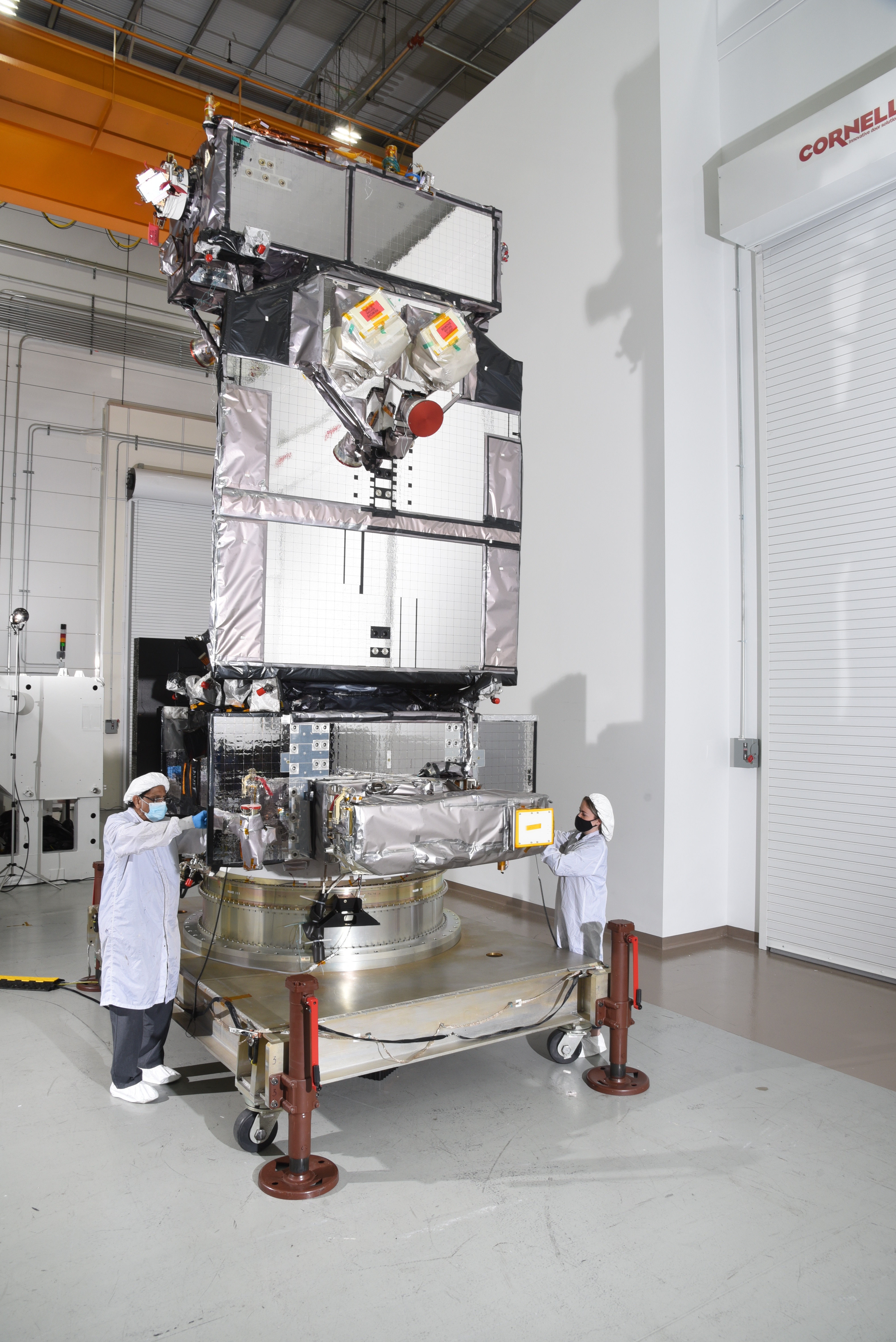A person wearing a white lab coat, black pants, a mask and gloves examines a large machine from the left and another person wearing a lab coast does the same from the right side.  
