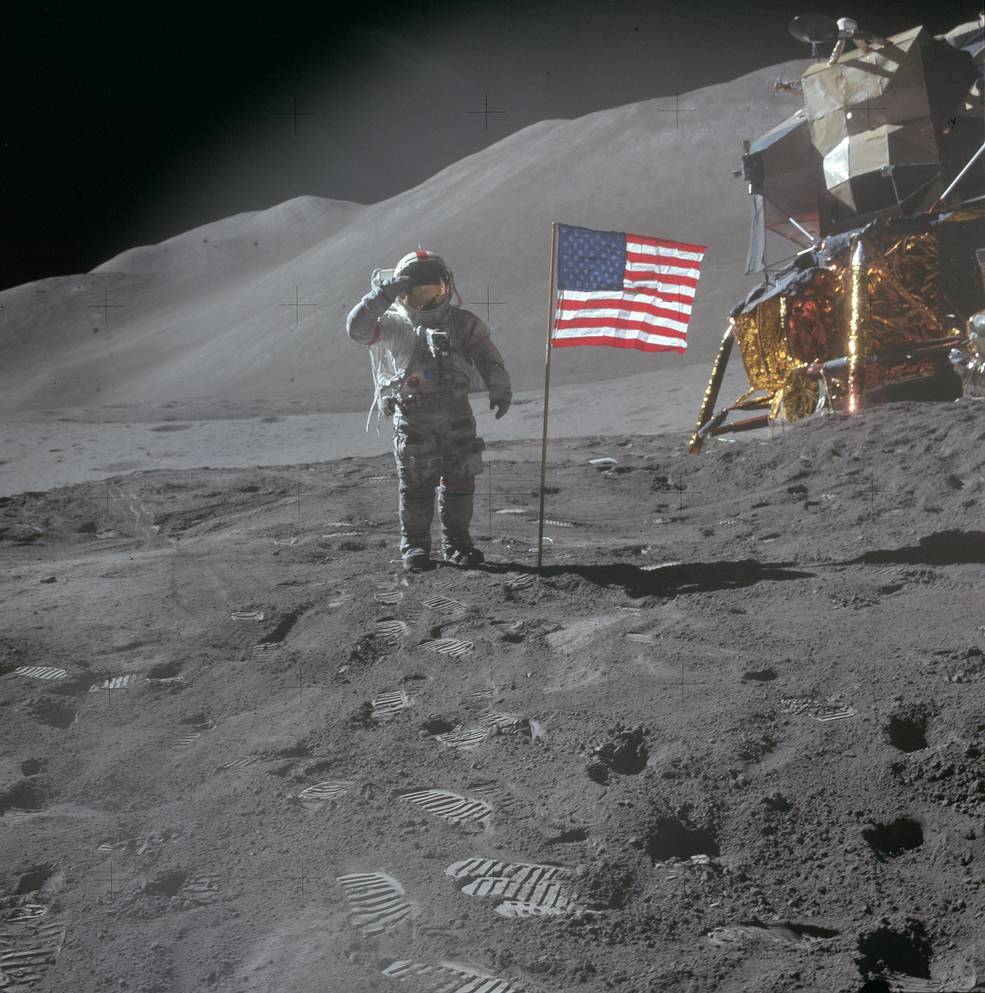An astronaut in a spacesuit stands on the lunar surface saluting the U.S. flag that's planted into the lunar surface. The lunar module is in the background along with some lunar hills at the Hadley-Apennine landing site.