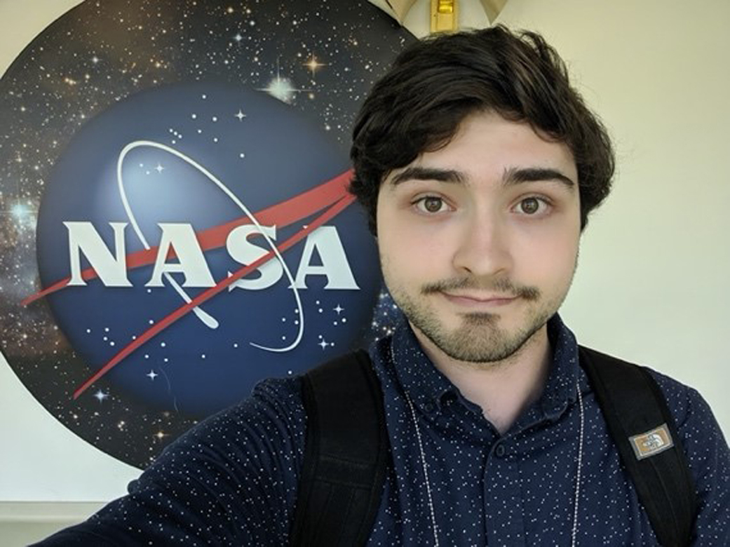 Young man with dark brown hair, black facial hair, and light tan skin softly smiles in front of a NASA meatball sign with stars behind it. He is wearing a navy collared shirt with a black backpack. 