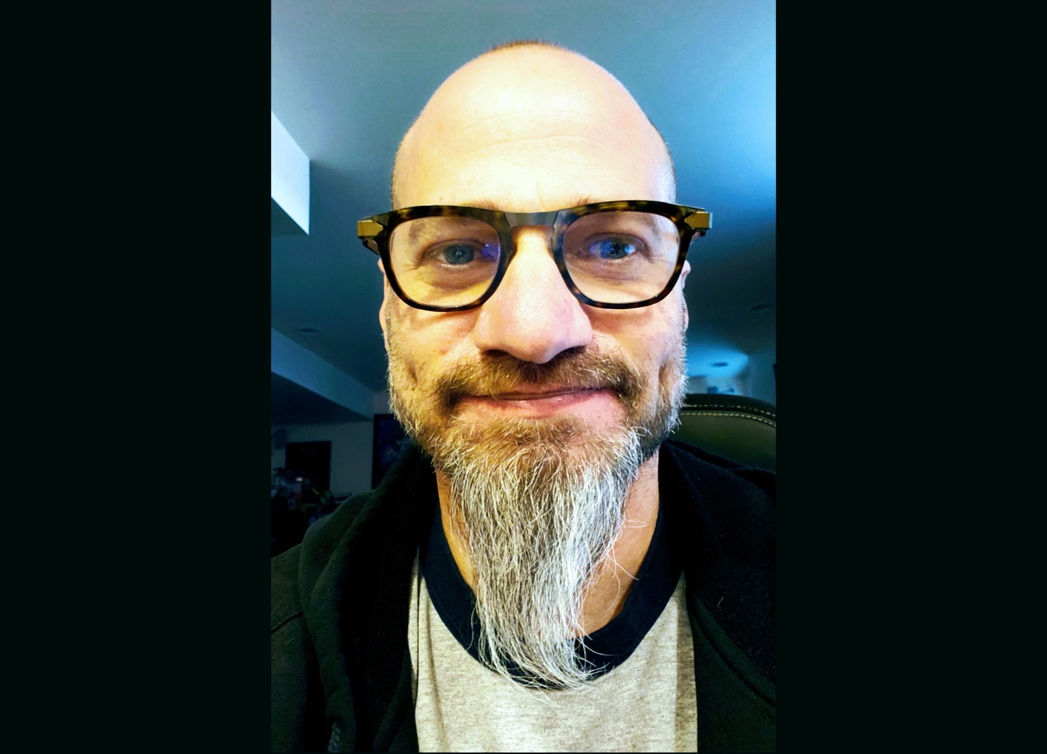 A man with light tan skin, a gray beard, little hair, and glasses wearing a gray shirt and jacket smiles at the camera