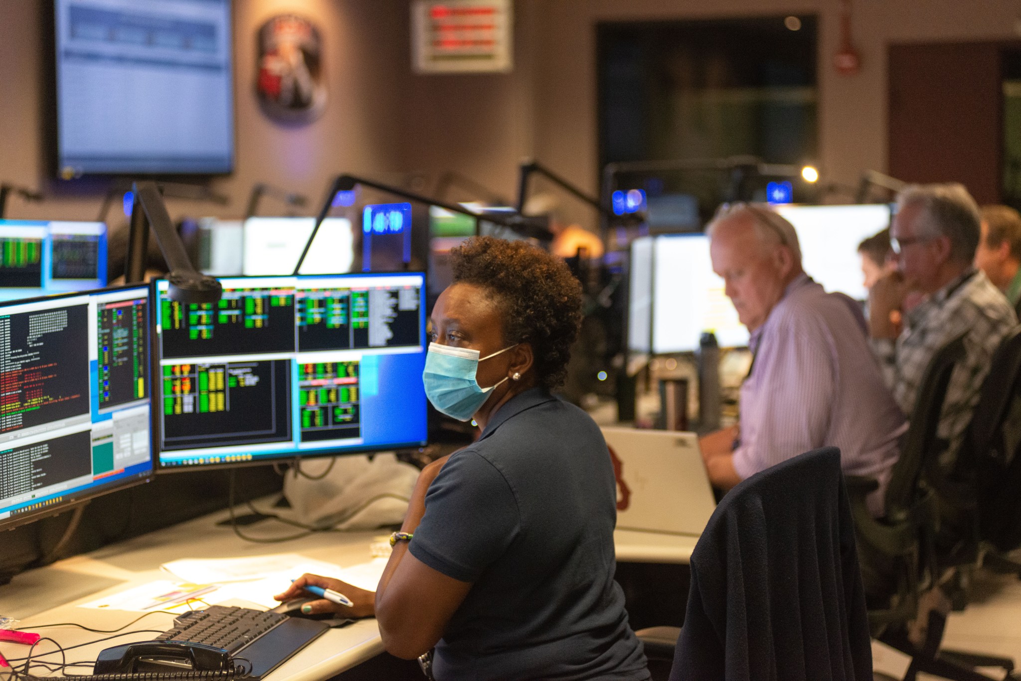Center: Nzinga Tull sits in front of several computer screens filled with computer code and data. To her right and farther back in the image sit several other Hubble Mission Operations Specialists.