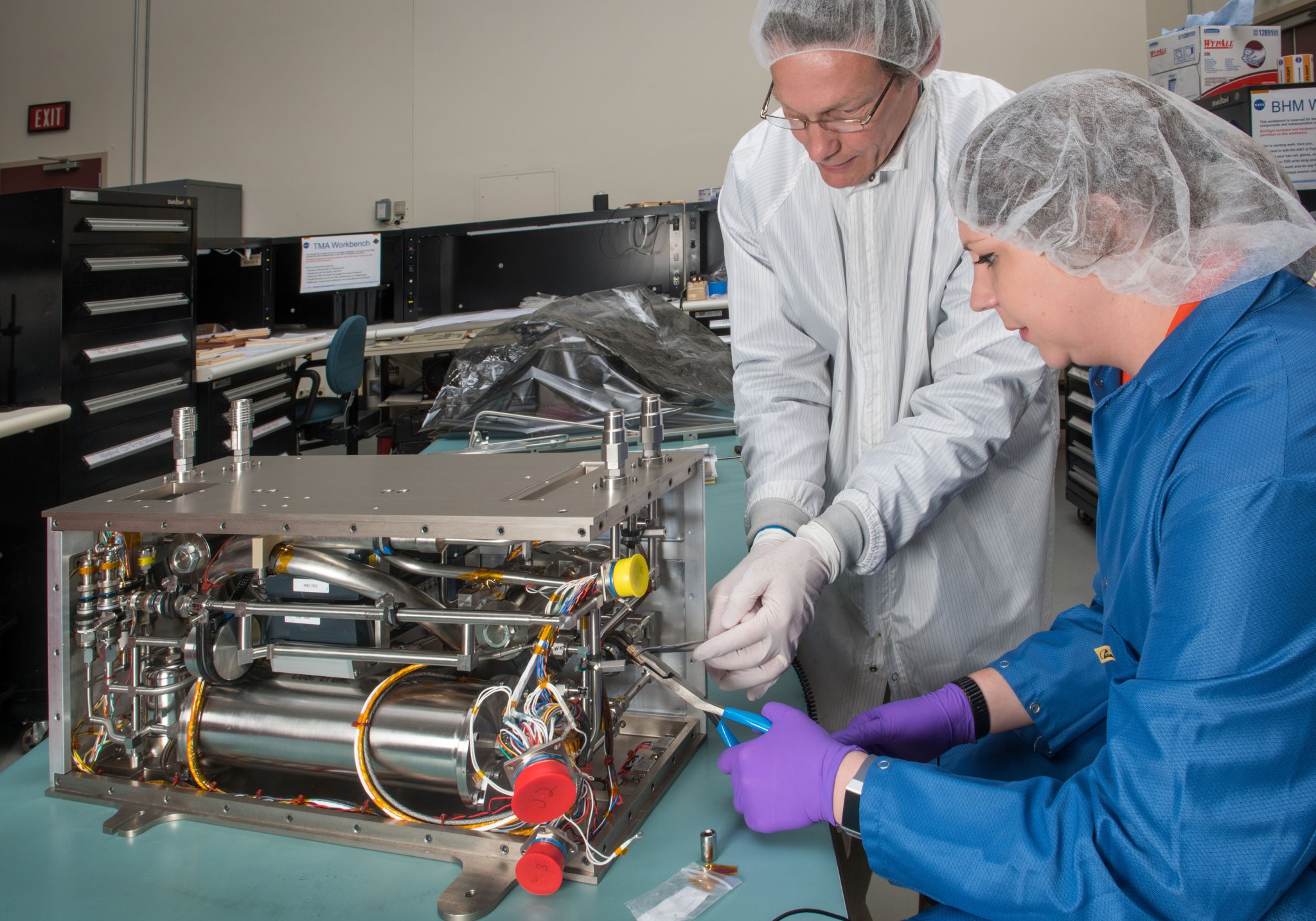 A man and woman in lab coats and hair nets work on space hardware.