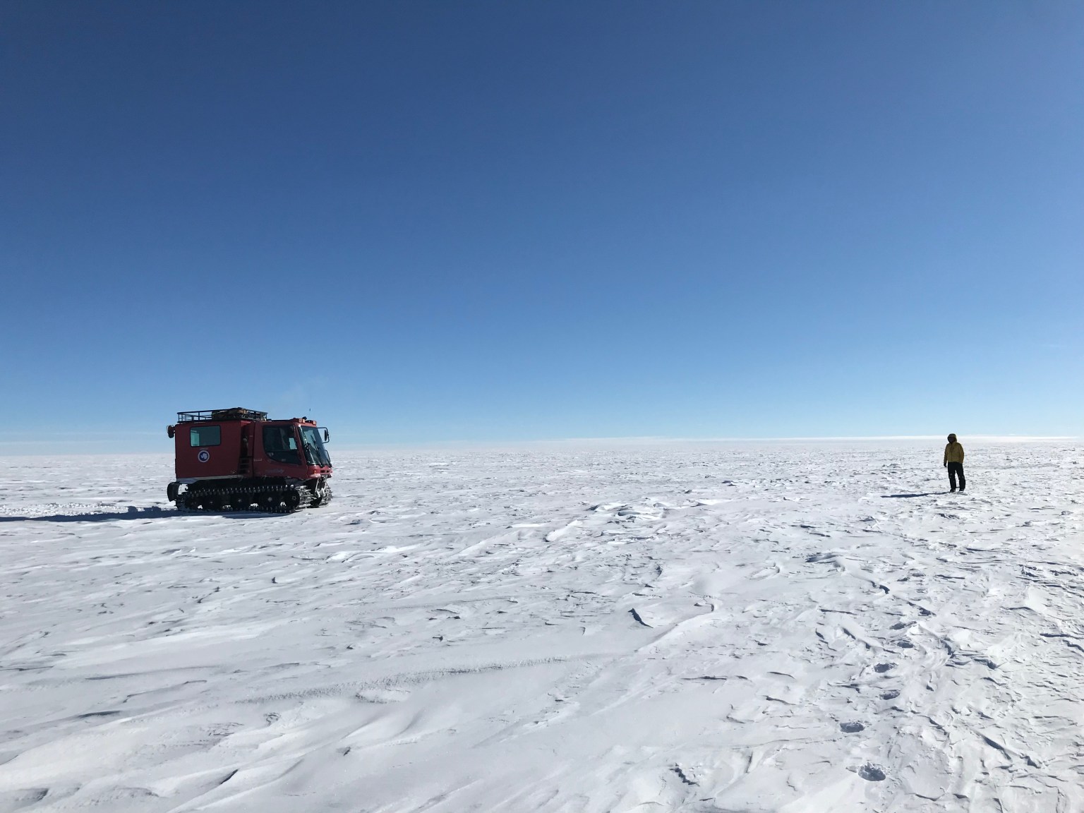 A scientist in winter clothes stands on a wide, flat, snow-covered plain with no trees or features, looking across at a boxy red snow vehicle with treads. The sky above them is cloudless and deep blue.
