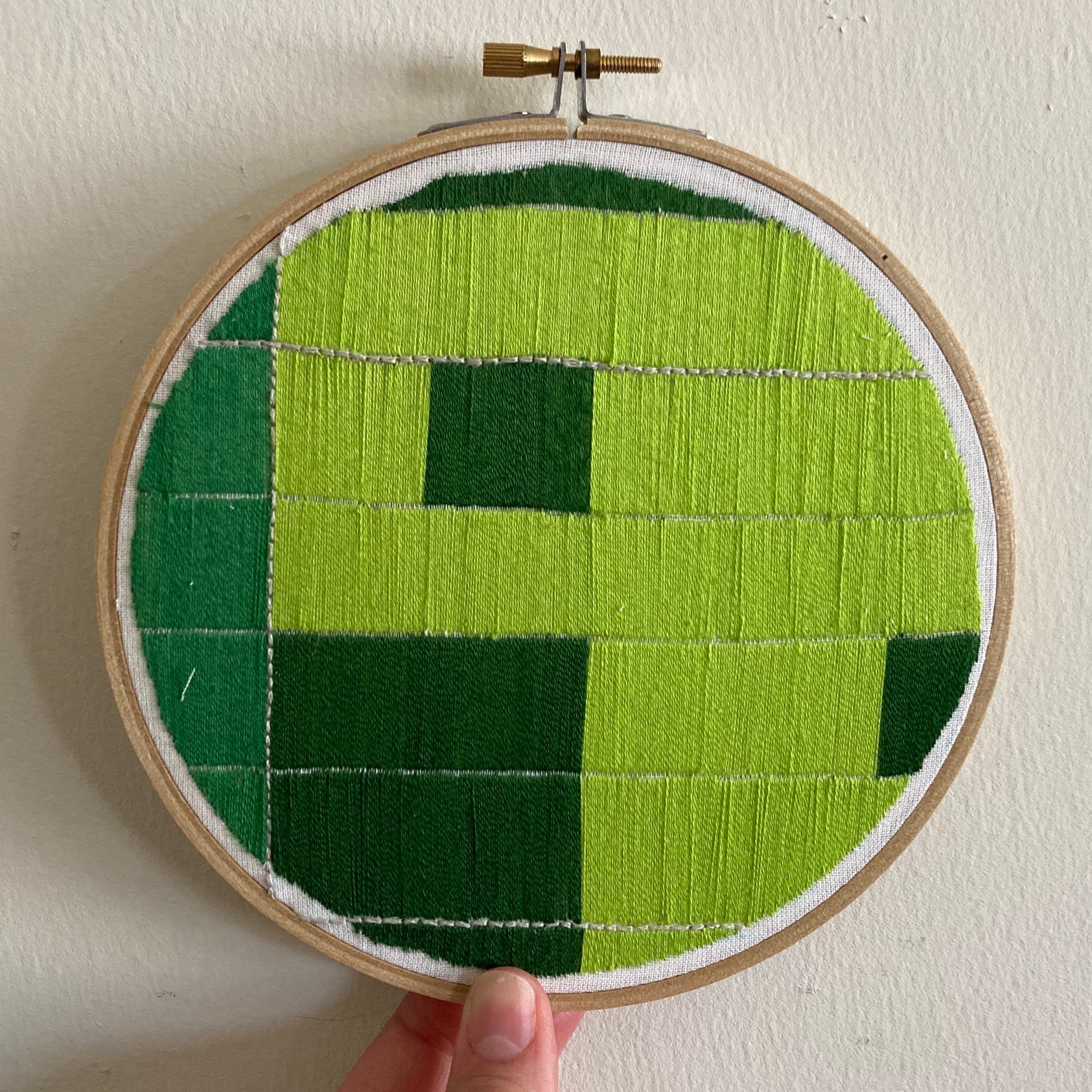 An embroidery hoop held in front of a white wall by two fingers. The embroidery is squares made of satin stitched straight lines, with three different shades of green, one color in each square.