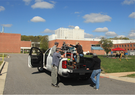 Four men load the hazar detection lidar system onto a metal rig and strapped it into the bed of a white pickup truck. The sky is blue with clouds and there are buildings at Goddard behind the truck. 