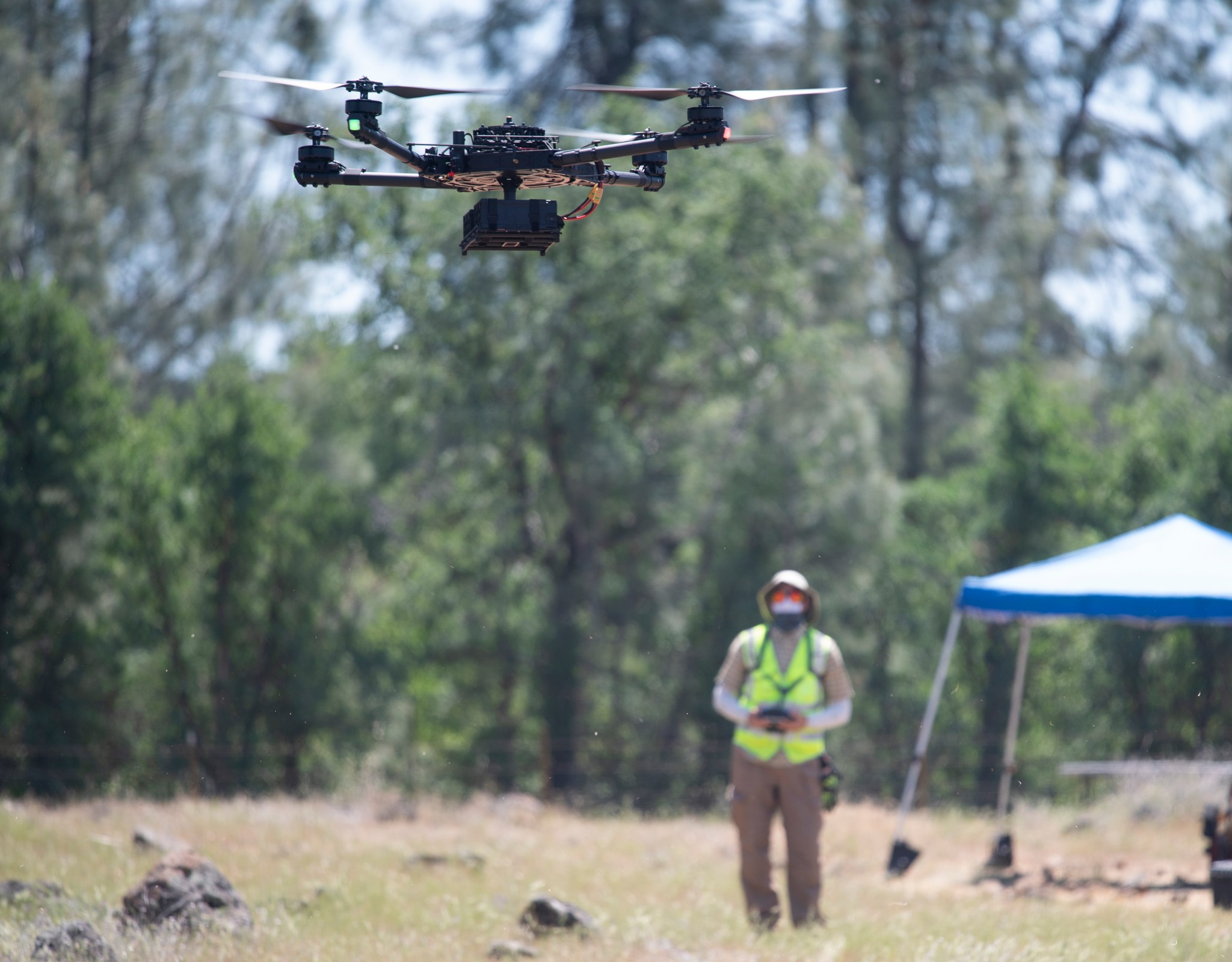 A quadcopter drone in flight in the foreground, with the pilot wearing a yellow vest in the background against a stand of trees.
