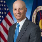 A white male in a suit poses for a picture in front of the American flag and the NASA flag