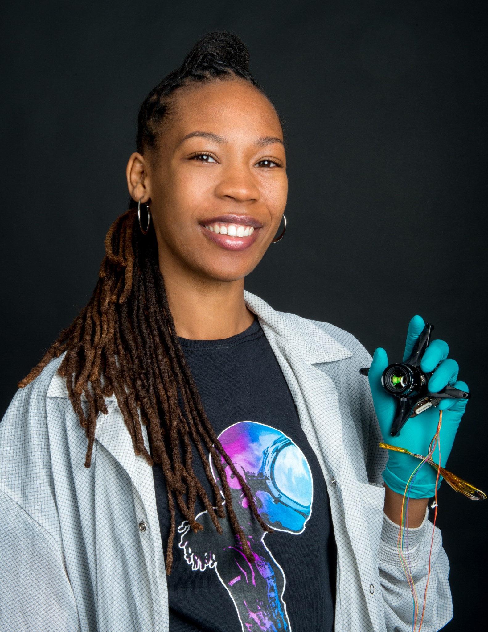 Sabrina Thompson wears a white lab coat over a shirt with graphic of a female astronaut. She is wearing blue gloves and holding a sensor in her hand.