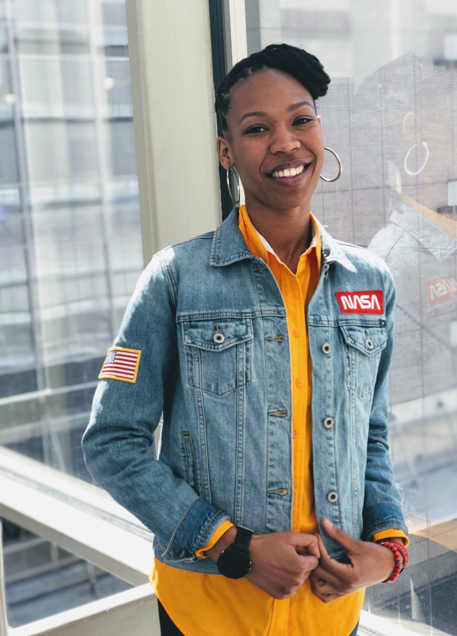 Sabrina Thompson standing in front of a window, smiling, wearing a yellow shirt with a jean jacket with a NASA patch on the right
