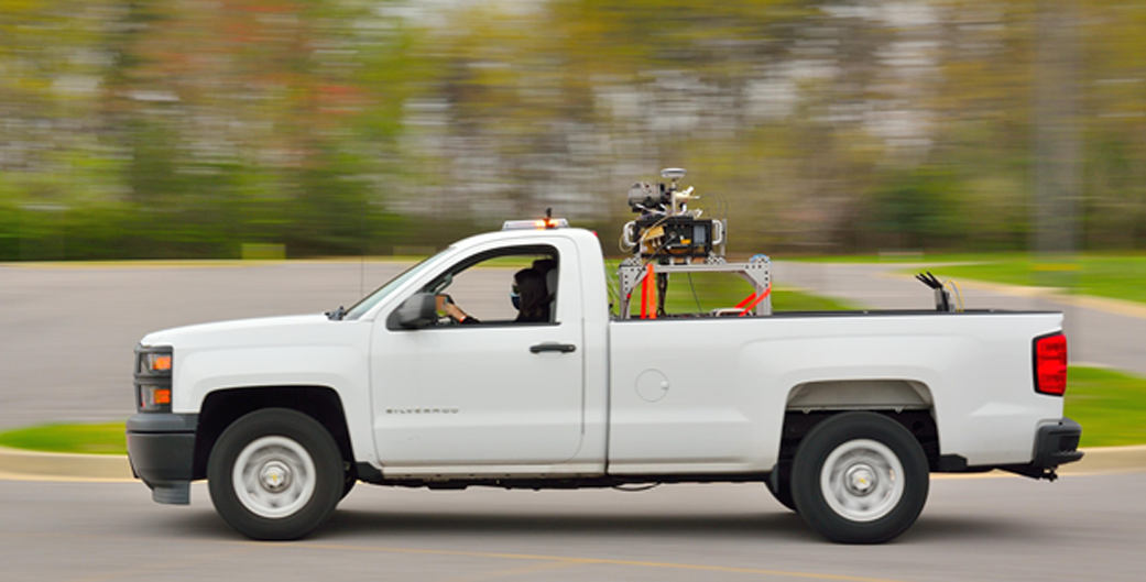 blurred background with trees and a white pickup truck in clear view. There's equipment in the back of the truck and a person driving