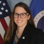 Image shows Zoe Sampson, an employee at NASA Kennedy posing for a photo wearing a black suit with the American flag and NASA flag behind her.