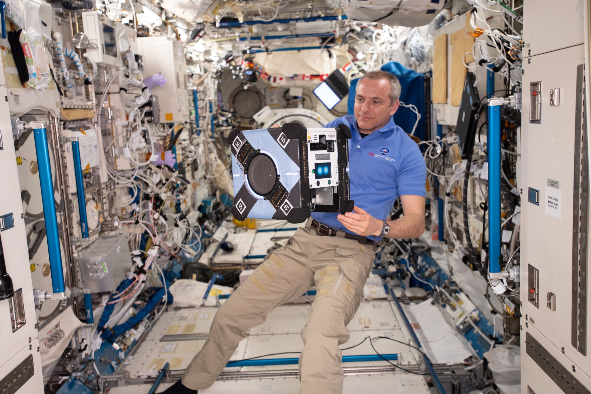 Man in a blue polo shirt floats next to a small, cube-shaped robot in a hallway in the International Space Station.