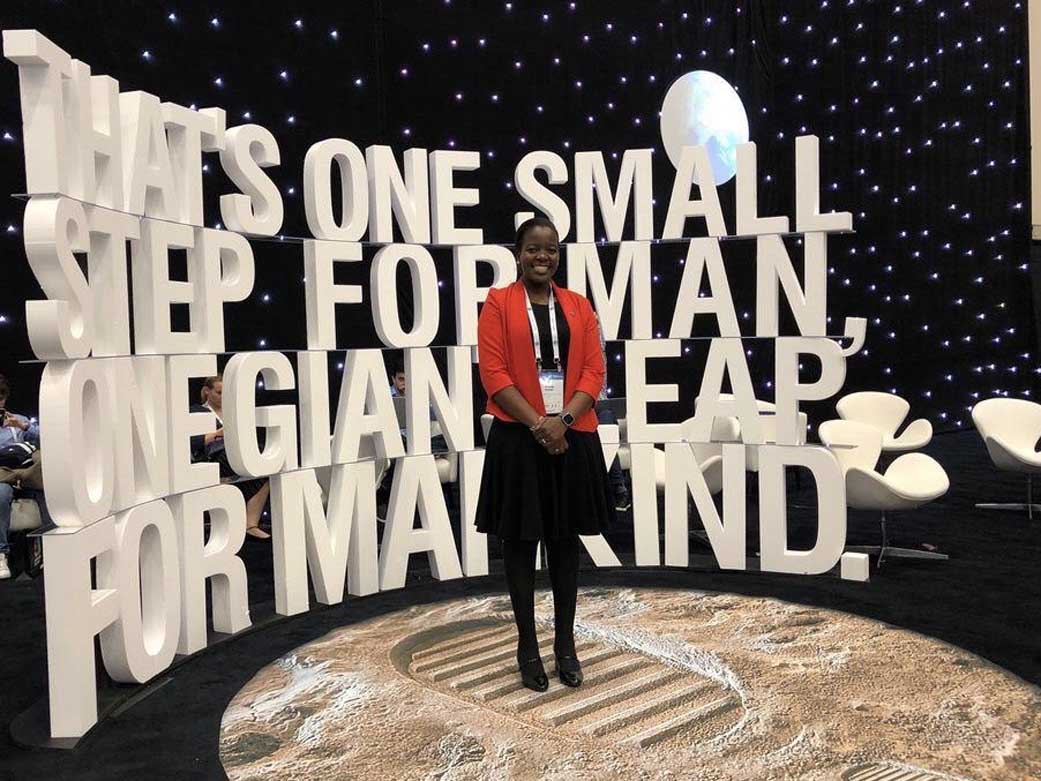 Dr. Mashiku stands on a large lunar footprint model in front of giant words that say "That's one small step for man, one giant leap for mankind." She is wearing a black dress, red blazer, black boots, and a white lanyard. 