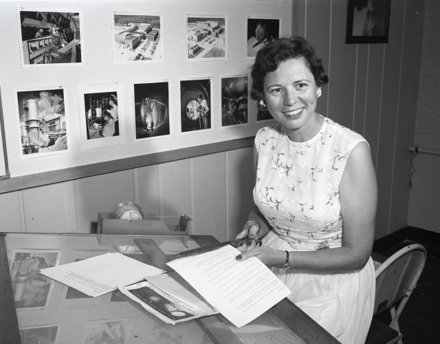 Woman smiling at camera holding scissors, cutting paper.