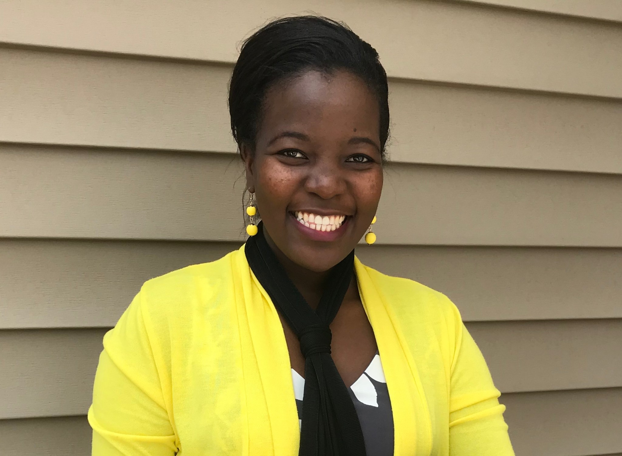 Dr. Alinda Mashiku, a Black woman, stands smiling against a tan wall. She is wearing a bright yellow sweater over a black and white shirt, a black scarf, and yellow earrings.