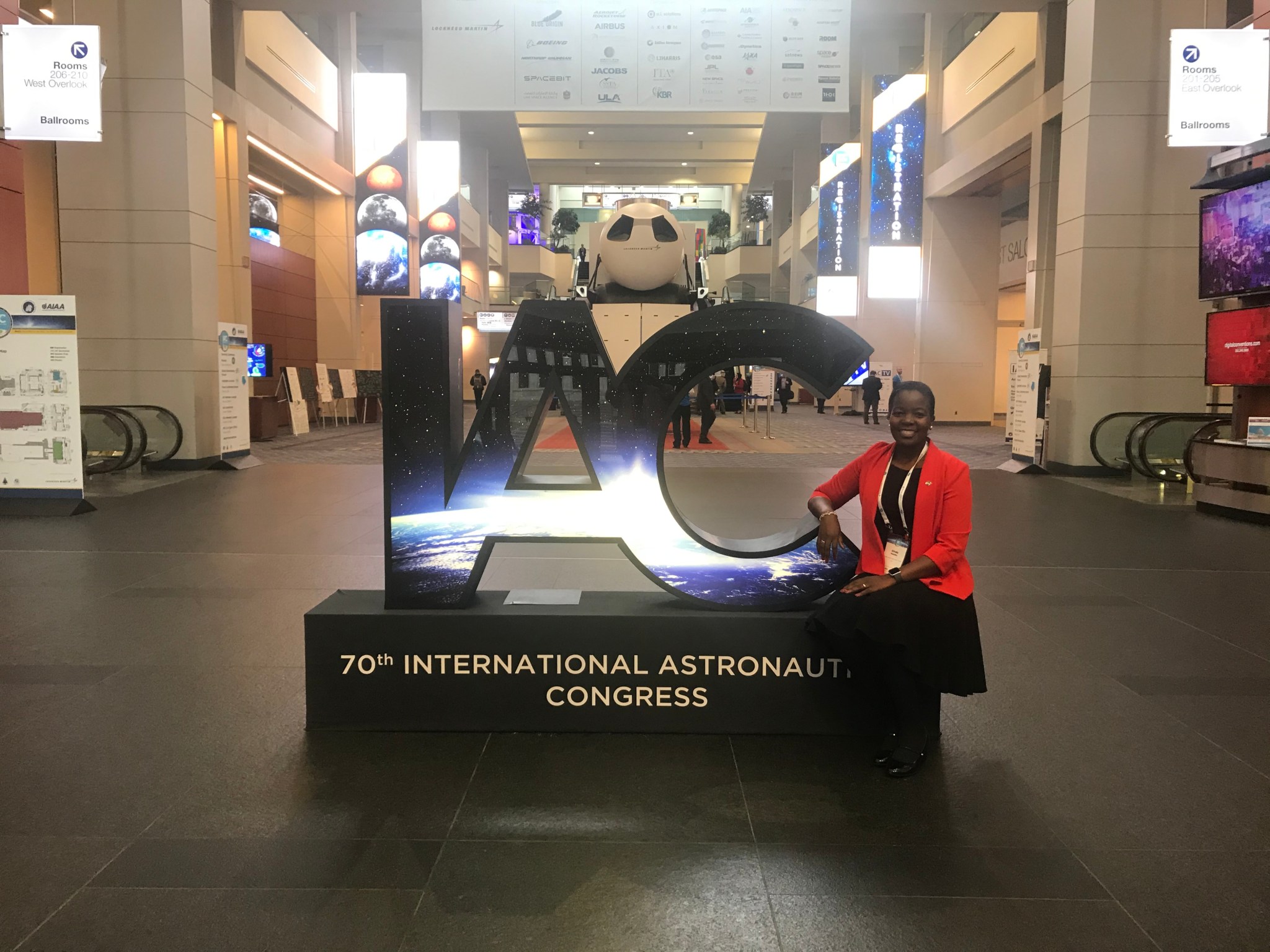 Dr. Mashiku sits on a sign with giant letters "IAC" and the words "70th International Astronaut Congress." She is wearing a black dress, red blazer, black boots, and a white lanyard. The background is a conference center.