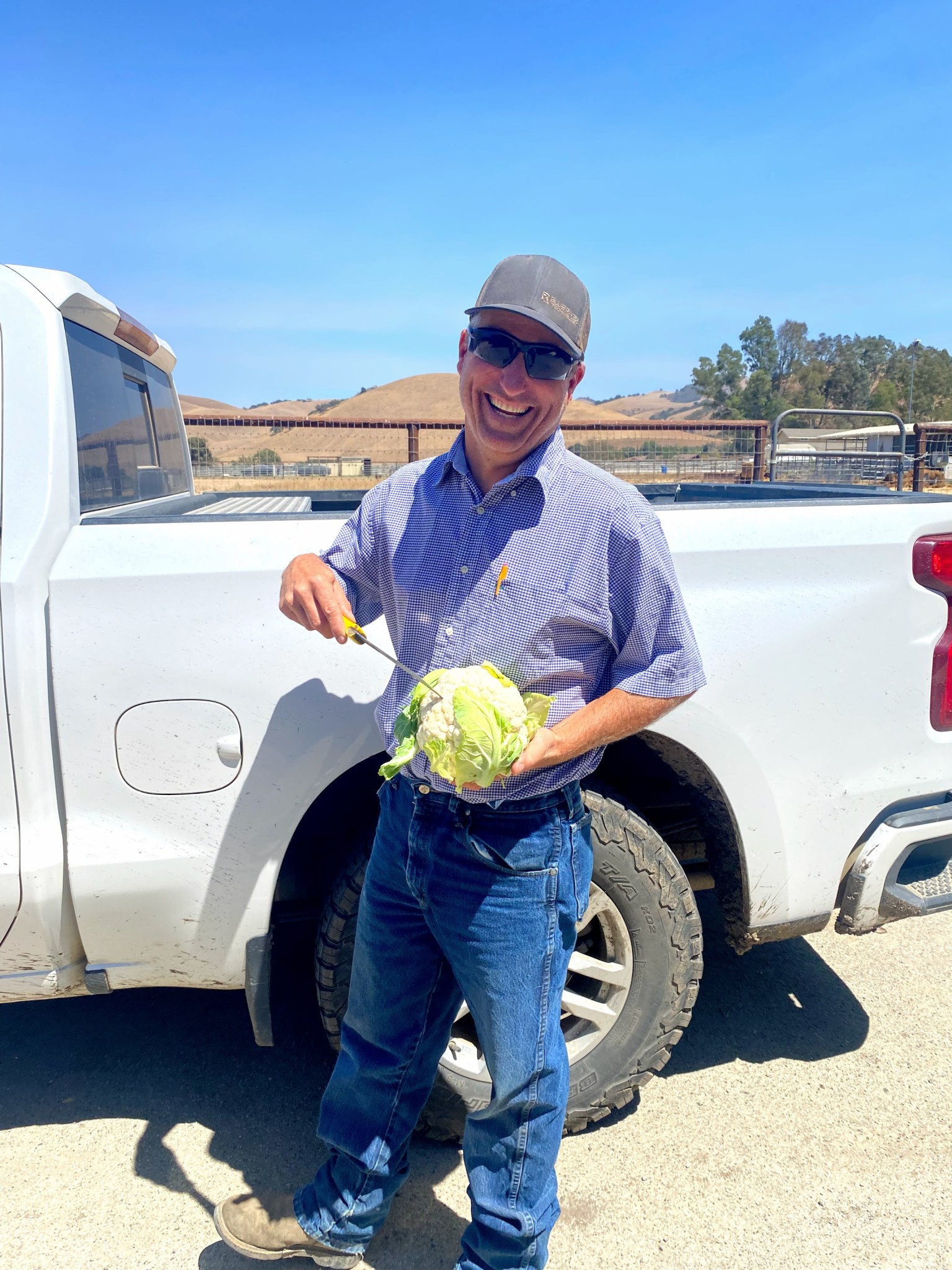 Farmer Mark Mason stands holding a knife and a head of cauliflower, smiling at the camera.