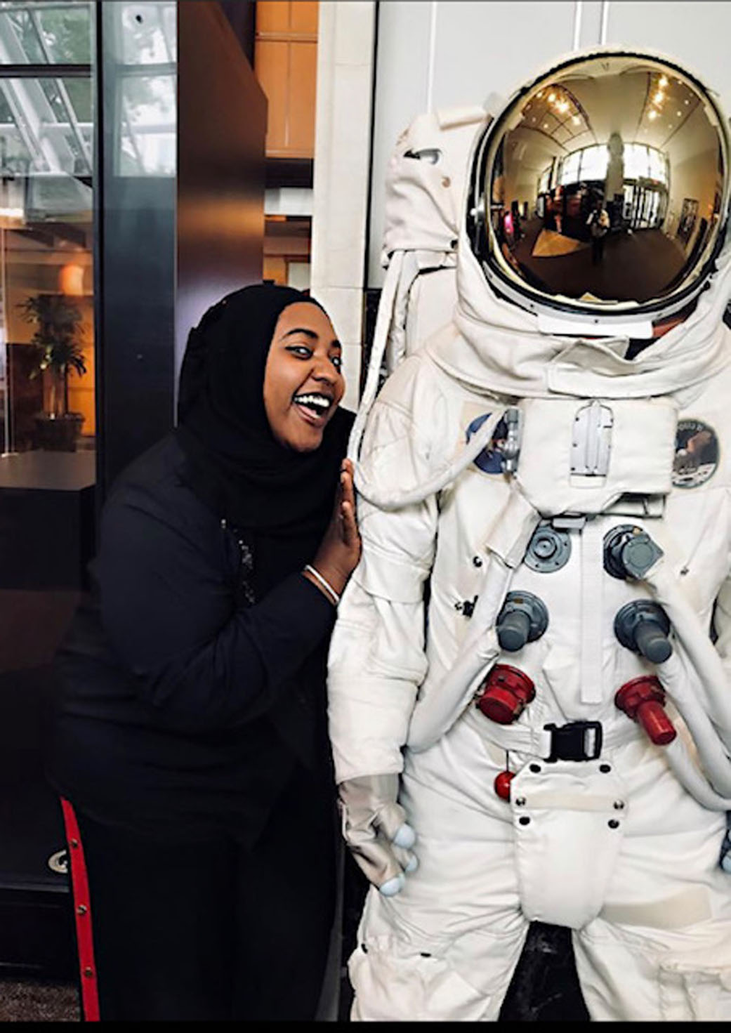 a Black woman wearing a black long-sleeved shirt and a black head covering smiles next to an astronaut suit. 