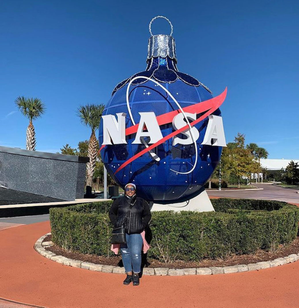 A Black woman wearing jeans, a black jacket, and sunglasses stands in front of an ornament shaped NASA meatball globe at Kennedy Space Center. 