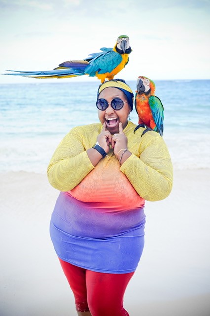 A Black woman wearing an ombre shirt of yellow, orange, purple, and blue with red leggings and black sunglasses stands in front of the ocean on a beach with two multicolored parrots. A blue and yellow parrot stands on her head and a red, green, and blue parrot sits on her shoulder. 