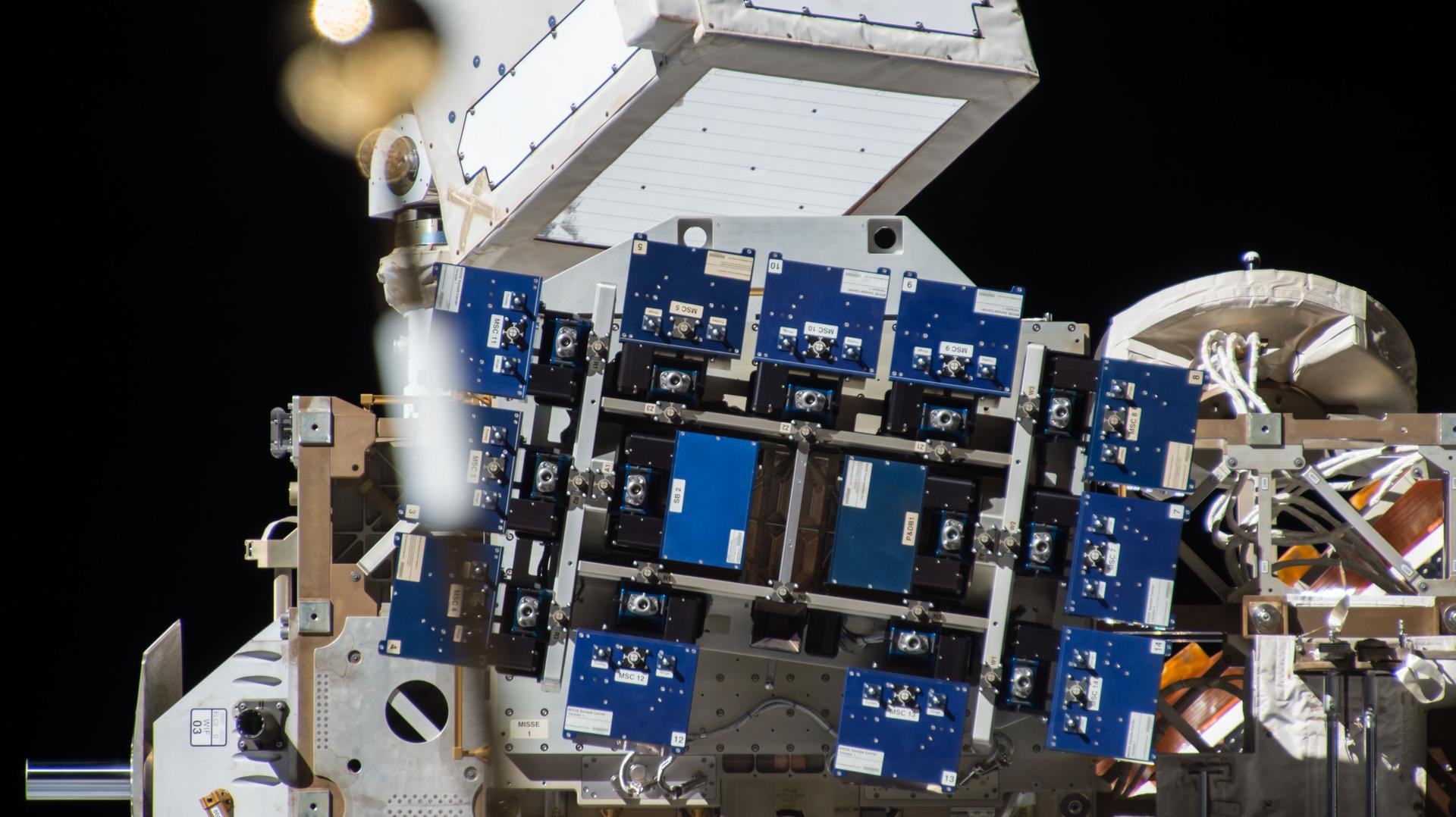 The MISSE-FF is visible in the image center, blue and black panels on a large white structure. The station's robotic arm extends from the top of the image and solar panels fill the background with the blackness of space behind them.