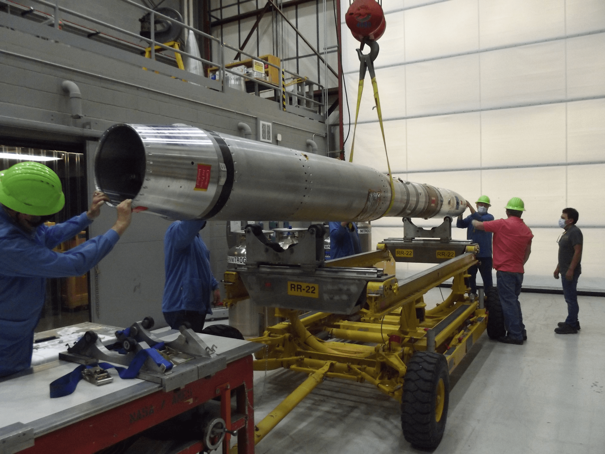 A crane lifts a silver rocket horizontally, while six individuals in hard hats guide the rocket onto a rolling cart.