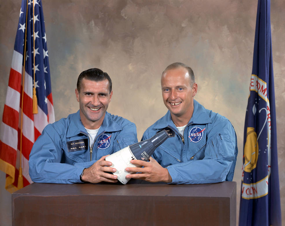 Two men posing with model space craft in front of flags forGemini XI crew photo