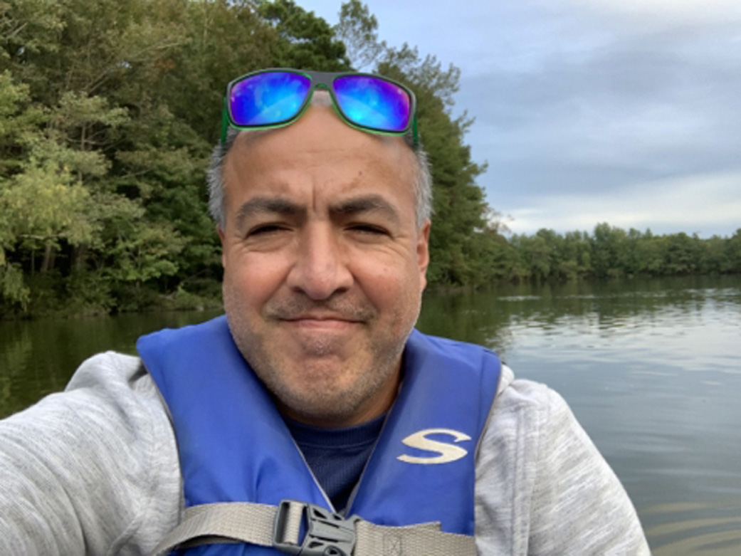 Man wearing a grey jacket, blue life jacket, and sun glasses on the top of his head, smiles with his mouth closed in a selfie. The man is sitting in a kayak with water surrounding him and trees along the edge of the water in the background. 