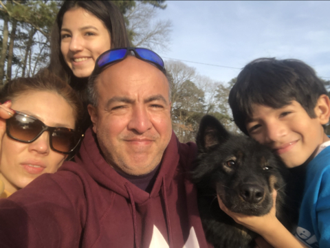 Selfie with four people and a dog in front of trees and a blue sky. From left to right, there's a woman wearing sunglasses, a young girl smiling, a man wearing a dark red hoodie and sun glasses on the top of his head, a black dog with pointed ears, and a young boy with black hair, wearing a blue shirt, holding the dog's face 