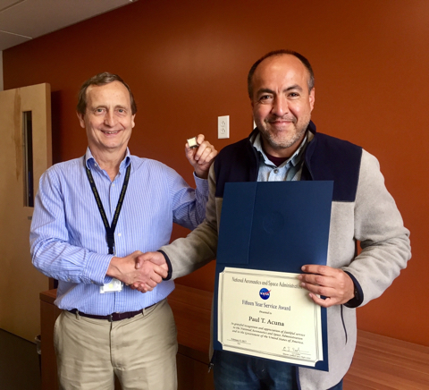 Man wearing a blue dress shirt and khaki pants shakes hands with a man wearing a tan and black jacket and jeans, holding his 15 year career award.