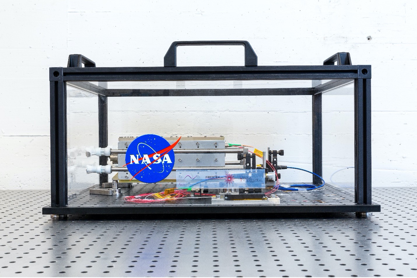 A laser prototype rests in a glass box on top of a perforated metal table.