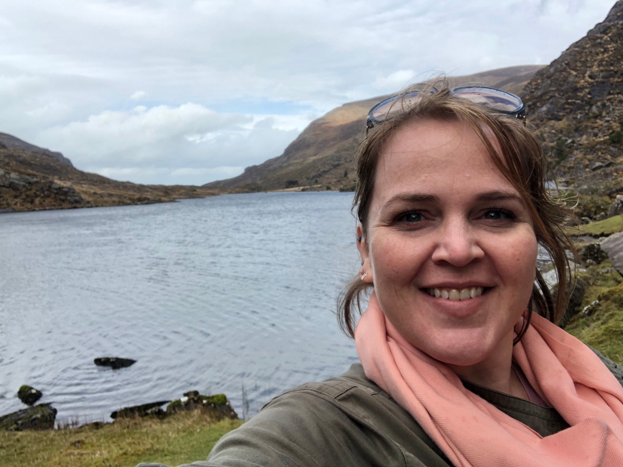 woman with brown hair in a ponytail wearing a coral scarf and dark green shirt stands smiling at the Gap of Dunloe in Ireland.