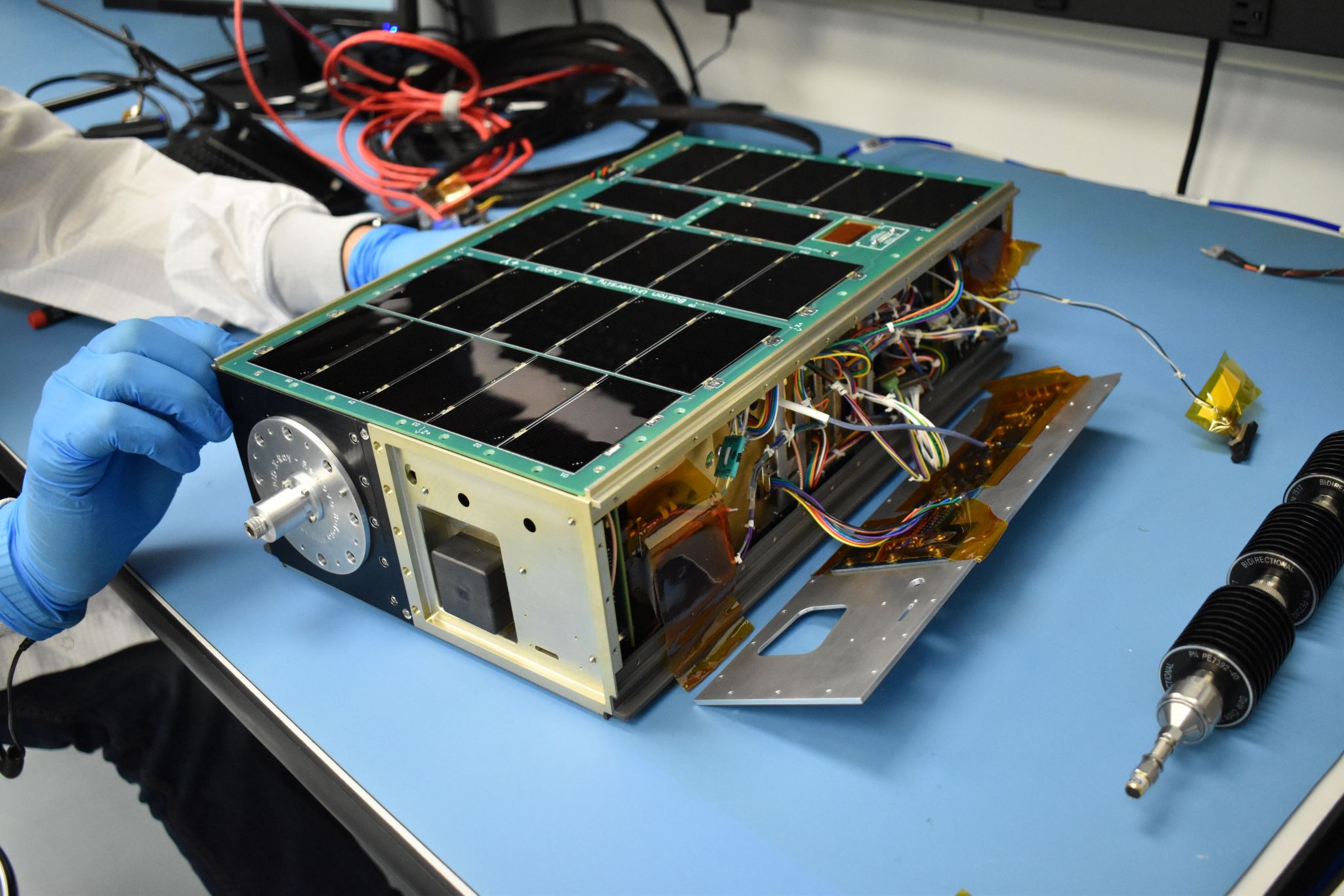 A cereal-box sized satellite with some wiring exposed sits on a table while someone with gloved hands works on it