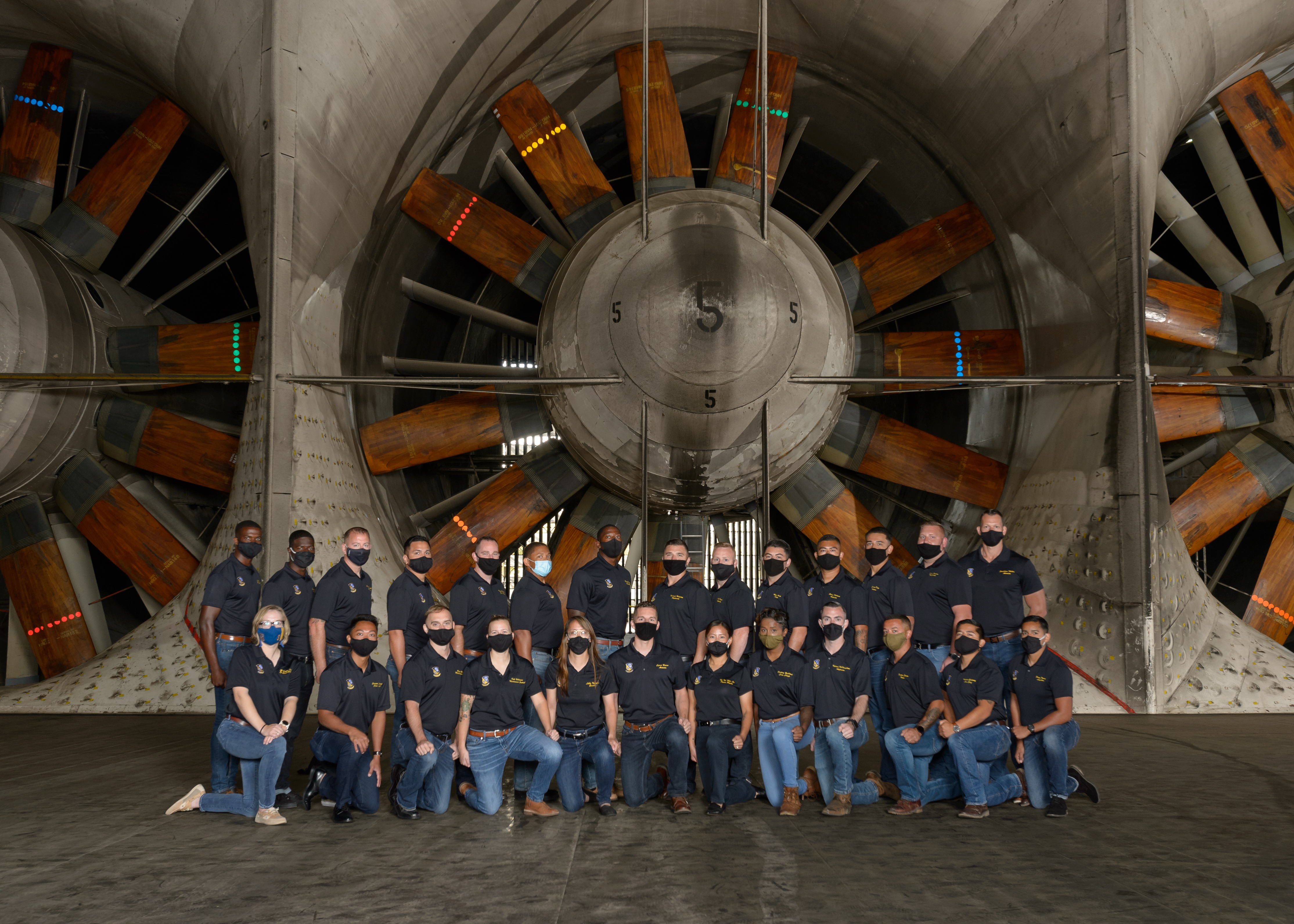 A large group of people all dressed in navy blue shirts and jeans pose in front of a fan 40 feet in diameter.