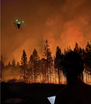 A drone with green lights flies against a background of wildfire in a forest. A figure in silhouette holds a tablet in the foreground.
