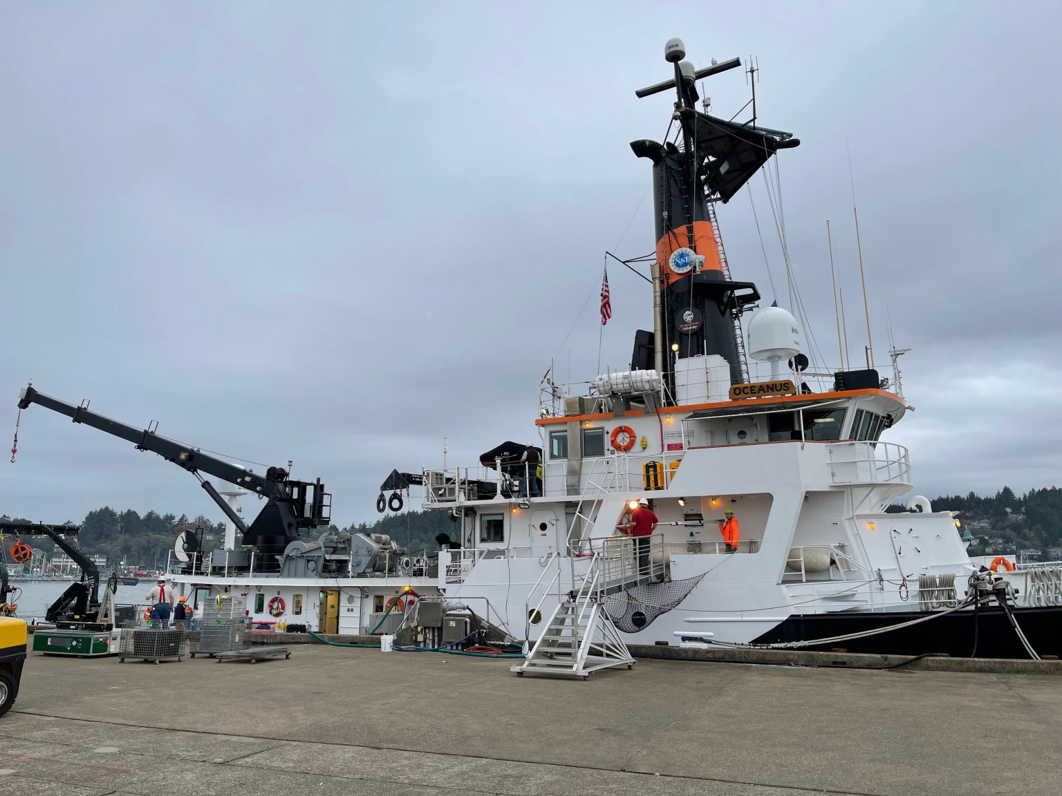 An image of a small ship at a dock before a science voyage. The ship is mostly white, with a black smokestack with an orange stripe. It has a black crane mounted on its back and a wooden plaque at the front of the bridge with its name, "Oceanus." A person wearing a bright orange jacket and yellow hardhat stands on one of the decks and looks across at the camera. It is a cloudy day, and heavy, dark clouds cover the sky.