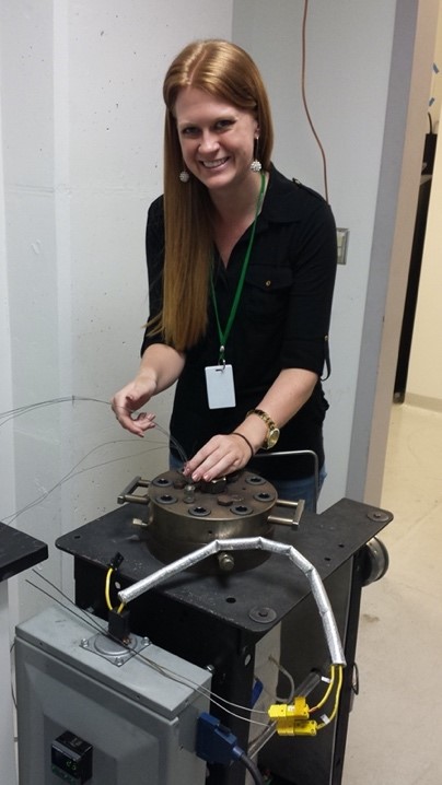 Woman with long red hair, wearing a black shirt, green lanyard, and white earrings works with a Venus chamber, a black machine with a round piece on top. 